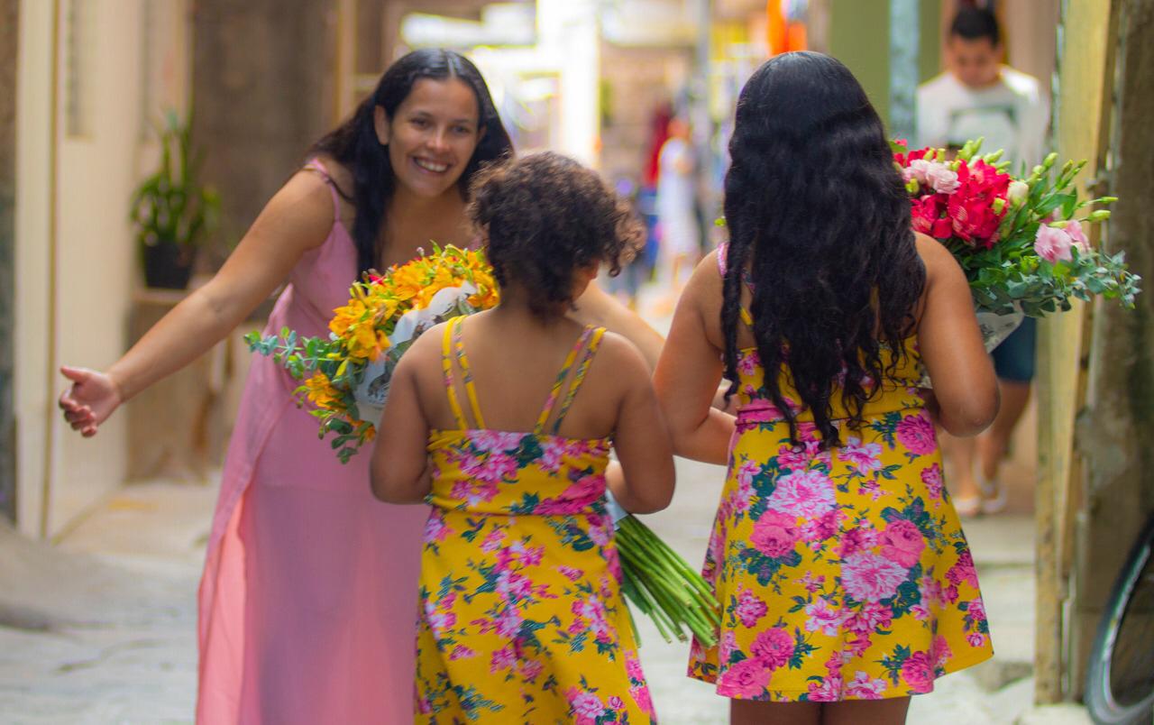 Mãe gestante com braços abertos para receber filhas pequenas com vestidos floridos 